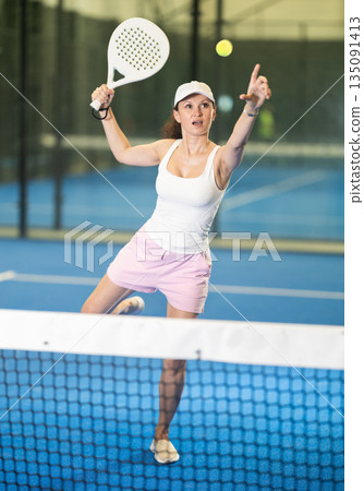Woman with racket training on indoor court playing padel game and hitting ball Woman with racket training on indoor court playing padel game and hitting ball 135091413