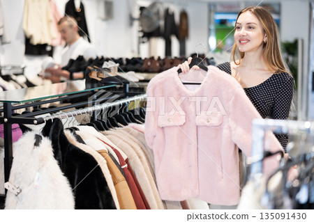 Girl stopped near showcase with outerwear, holds fur coat hanger in hands and examines product Girl stopped near showcase with outerwear, holds fur coat hanger in hands and examines product 135091430