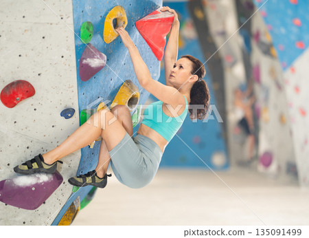 Adult woman is mastering climbing on training wall in gym, side view. Young woman holds on tightly to ledges and strives for top of bouldering route 135091499