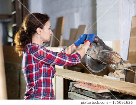 Female employee of workshop cuts wooden board into narrow parts using circular saw Female employee of workshop cuts wooden board into narrow parts using circular saw 135091626