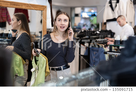 While shopping, girl looks at blouse and chats on phone with friend, discusses latest news. While shopping, girl looks at blouse and chats on phone with friend, discusses latest news. 135091715