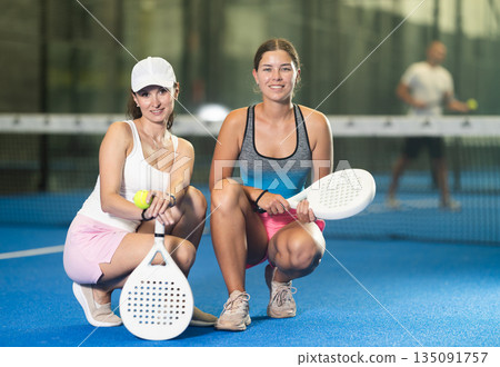 Smiling sportswomen standing next to the net with padel racket and tennis ball in hands in tennis court Smiling sportswomen standing next to the net with padel racket and tennis ball in hands in tennis court 135091757