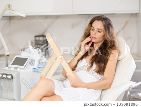 Positive woman examining her face after cosmetic procedure in medical room 135091896