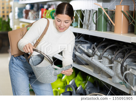 Amateur gardener girl selects watering can for her hobby in greenhouse flower-bed 135091905
