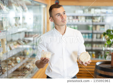 Attentive young man purchaser choosing cheese in large supermarket Attentive young man purchaser choosing cheese in large supermarket 135091961