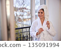 Healthy lifestyle, hair maintenance, morning beauty. Woman in white bathrobe happily dries her hair with towel near window overlooking serenity. 135091994