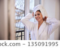 Serene background, window light, morning routine. Woman in white bathrobe happily towels her hair dry while standing near serene window. 135091995
