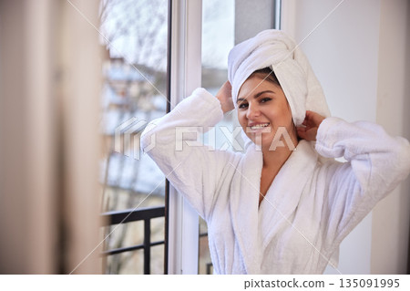 Serene background, window light, morning routine. Woman in white bathrobe happily towels her hair dry while standing near serene window. Serene background, window light, morning routine. Woman in white bathrobe happily towels her hair dry while standing near serene window. 135091995
