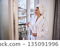 White bathrobe, Peaceful moment, Looking outside. Wearing white bathrobe and towel wrap, woman stands by balcony, peacefully gazing at scenery outside. 135091996