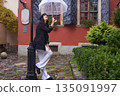 Fashionable woman, Woman outdoors, Rain clothing. In stylish black coat and beret, woman smiles while walking on rainy cobblestone street, holding transparent umbrella. 135091997