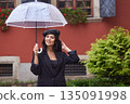 Umbrella rain, Rain accessories, Stylish coat. During rainy day, woman dressed in fashionable black coat and beret holds clear umbrella and smiles while walking on cobblestone street. 135091998