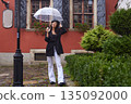 Cobblestone street, Rain fashion, Umbrella rain. On rainy day, woman in stylish black coat and beret smiles while holding transparent umbrella and strolling along cobblestone street. 135092000