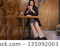 Woman coffee, Cafe table, Rustic decor. Woman enjoys coffee while seated at cafe table, surrounded by rustic wheat-themed decor, creating warm and inviting atmosphere. 135092003