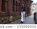 Casual smile, city stroll, fashion photography. Stylish woman walks past brick wall on rainy day, wearing black jacket and white pants and smiling casually. 135092010