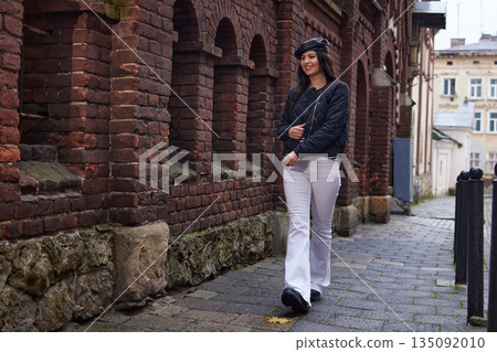 Casual smile, city stroll, fashion photography. Stylish woman walks past brick wall on rainy day, wearing black jacket and white pants and smiling casually. 135092010