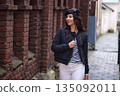 Black jacket, autumn stroll, black coat. Woman wearing black jacket and beret smiles as she walks along cobblestone street beside old brick wall in autumn season. 135092011