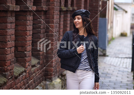 Black jacket, autumn stroll, black coat. Woman wearing black jacket and beret smiles as she walks along cobblestone street beside old brick wall in autumn season. 135092011