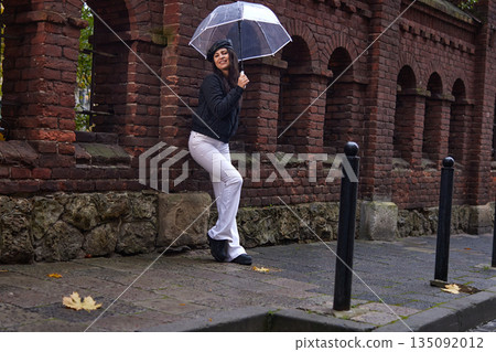 Clear umbrella, city rain, fashion portrait. Confident, smiling woman holds transparent umbrella as she leans against aged brick wall on rainy day. Clear umbrella, city rain, fashion portrait. Confident, smiling woman holds transparent umbrella as she leans against aged brick wall on rainy day. 135092012