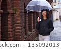 Rainy day, white pants, brick wall. Strolling beside old brick wall on rainy day, stylish woman in black quilted jacket and white pants smiles under clear umbrella. 135092013