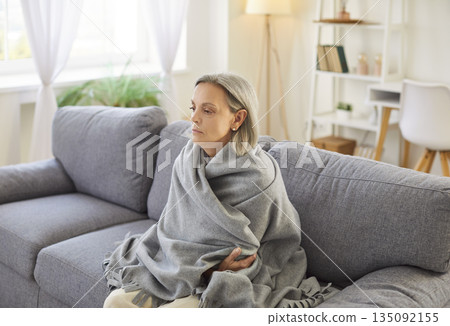 Freezing elderly woman sitting wrapped in plaid, trying to warm up, suffering from low temperature. 135092155