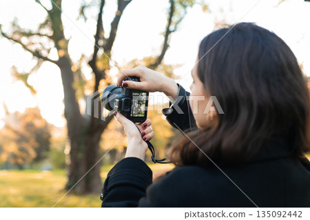 Young woman reviewing photos on her digital camera while standing in a sunny park. Warm autumn light, creative outdoor photography moment, lifestyle scene. Young woman reviewing photos on her digital camera while standing in a sunny park. Warm autumn light, creative outdoor photography moment, lifestyle scene. 135092442