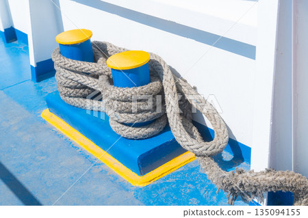 A detailed view of a thick rope tied around a blue bollard, located on a boat near a calm water body. Represents maritime activities, boating and nautical themes. High quality photo 135094155