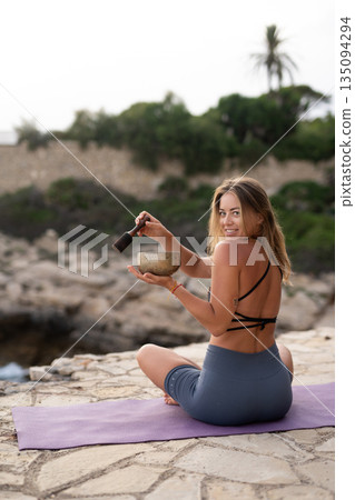 Female yoga practitioner relaxing on a mat by the coast, using a sound healing bowl to create a calm and spiritual atmosphere. 135094294