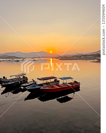Serene sunset over calm water with moored boats and distant mountain 135094604
