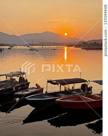 Serene sunset over calm water with moored boats and distant mountains 135094606