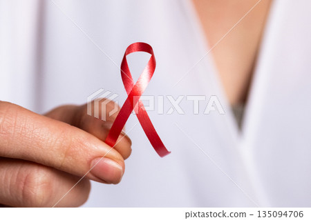 A doctor holds a red ribbon in their hand, symbolizing awareness for World Cancer Day on February 4th. The doctor wears a white coat and shows commitment to cancer awareness. 135094706