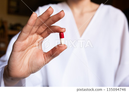 A young woman doctor in a white coat holds a red and white capsule in her right hand. The background is softly blurred, emphasizing the pill and the doctor. A young woman doctor in a white coat holds a red and white capsule in her right hand. The background is softly blurred, emphasizing the pill and the doctor. 135094746