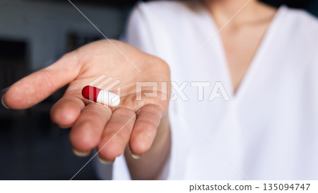 A young woman doctor in a white coat holds a red and white capsule in her right hand. The background is softly blurred, emphasizing the pill and the doctor. 135094747