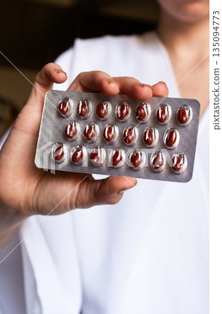 A female doctor holds a blister pack of red pills. She wears a white coat and has long dark hair. The background is softly blurred, focusing on the medication. A female doctor holds a blister pack of red pills. She wears a white coat and has long dark hair. The background is softly blurred, focusing on the medication. 135094773