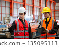 Two industrial workers in safety vests and helmets discussing operations while reviewing checklist on clipboard inside factory. Concept of team communication, planning and workplace safety. 135095113