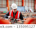 Industrial engineer wearing white safety helmet and red reflective vest operates heavy machinery in manufacturing plant, emphasizing focus, precision and hands on work in heavy factory. 135095127