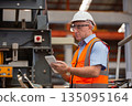 Experienced factory engineer in safety vest and helmet using tablet to inspect industrial equipment at production plant. Concept of digital technology in modern manufacturing operations. 135095164