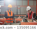 Two factory workers in safety uniform inspecting machinery with clipboard in manufacturing plant. Industrial teamwork concept focusing on engineering process, precision and maintenance. 135095166