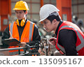Two factory workers in safety uniform inspecting machinery with clipboard in manufacturing plant. Industrial teamwork concept focusing on engineering process, precision and maintenance. 135095167