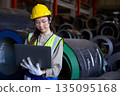Female engineer inspects data on laptop inside metal sheet factory warehouse, focusing on production monitoring, inventory tracking and industrial workflow efficiency. 135095168