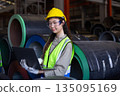 Female engineer inspects data on laptop inside metal sheet factory warehouse, focusing on production monitoring, inventory tracking and industrial workflow efficiency. 135095169