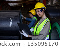 Female engineer inspects data on laptop inside metal sheet factory warehouse, focusing on production monitoring, inventory tracking and industrial workflow efficiency. 135095170