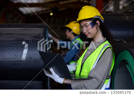 Female engineer inspects data on laptop inside metal sheet factory warehouse, focusing on production monitoring, inventory tracking and industrial workflow efficiency. Female engineer inspects data on laptop inside metal sheet factory warehouse, focusing on production monitoring, inventory tracking and industrial workflow efficiency. 135095170