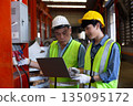 Two industrial engineers wearing safety helmets and reflective vests work together in factory using laptop, focusing on electrical system inspection, data analysis, and equipment monitoring. 135095172