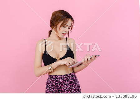 Young women in black crop top and floral skirt using digital tablet on pastel pink background, representing modern lifestyle, technology, fashion and online communication. 135095190