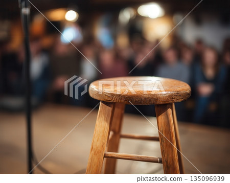 Wooden stool and microphone stand on a stage with a blurred audience watching, emphasizing anticipation, performance, stand up comedy, and live entertainment in a club setting 135096939