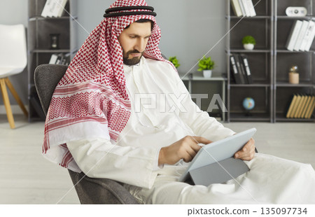 Business professional in keffiyeh using digital tablet while seated in office 135097734