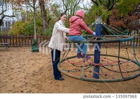 Mother Steadying Daughter On Spinning Roundabout Active Play With Supervision, Autumn Park, Jacket And Hat, Trusting Grip, Energetic Circle Of Motion 135097968
