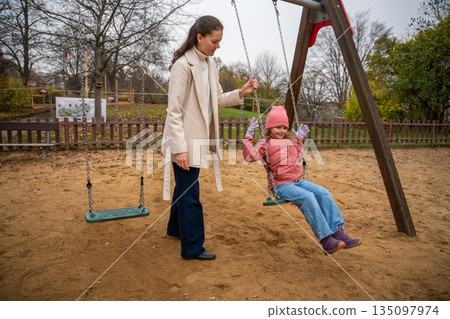 Smiling Child On Swing With Mother In Autumn Park Smiles Brightly, Mother Pushes Her Daughter On Green Swing Amid Autumn Trees And Sandy Ground Smiling Child On Swing With Mother In Autumn Park Smiles Brightly, Mother Pushes Her Daughter On Green Swing Amid Autumn Trees And Sandy Ground 135097974