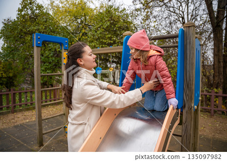Mother Guiding Daughter Down Playground Slide Careful Hands On Jacket, Shiny Metal Slide, Autumn Leaves, Supportive Posture, Smiling Trustful Moment 135097982