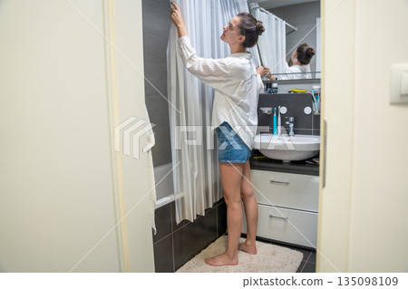 A woman standing barefoot in a modern bathroom adjusts a shower curtain near the bathtub and sink. Real life home interior scene showing everyday domestic routine, hygiene habits and home maintenance. 135098109